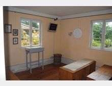 Ein kleiner Ausstellungsraum mit zwei großen Fenstern, die einen Blick auf einen Garten mit blühenden Pflanzen bieten. Im Raum befinden sich Holzfußboden, eine Glasvitrine auf einem schmalen Tisch, Bilder an der Wand, ein Holzfass und eine Sitzbank mit Marmorsitzfläche.