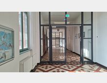 Langer Innenflur eines öffentlichen Gebäudes mit weiß gestrichenen Wänden, großen Fenstern links und einem gemusterten Fliesenboden in Rot- und Beigetönen. Im Vordergrund stehen schwarze Glasrahmentüren mit Fluchtweg-Schild, entlang des Flurs sind Heizkörper, Türen und gerahmte Bilder an der Wand zu sehen.