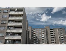 Wohnblock aus Betonplatten mit mehreren Etagen, Balkonen und Satellitenschüsseln, im Hintergrund ein bewölkter blauer Himmel. Die Fassade zeigt typische Gestaltungselemente von Plattenbauten, wie strukturierte Balkonbrüstungen.