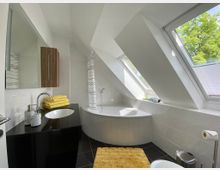 Helles, modernes Badezimmer mit Dachschrägen und großen Dachfenstern. Es verfügt über eine Eckbadewanne, ein schwarzes Waschbecken mit gelben Handtüchern darauf und einen weißen Fliesenboden mit einem gelben Teppich.