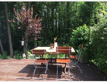 Holzterrasse mit einem Tisch und vier Metallstühlen mit Holzlatten, umgeben von dichtem Wald und Sträuchern; im Vordergrund ein kleiner Baum mit rötlichen Blättern.