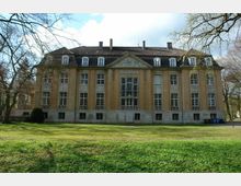 Historisches Gebäude mit symmetrischer Fassade, hohen Fenstern und einem steilen Dach, umgeben von einem Rasen und einzelnen Bäumen. Im Hintergrund ist ein bewölkter Himmel sichtbar.