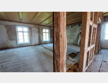 Ein unfertiger Innenraum in einem Altbau mit freiliegenden Holzträgern und unverputzten Wänden. Sichtbar sind Holzfußböden, zwei große Fenster mit weißem Rahmen sowie Deckenbalken aus Holz.