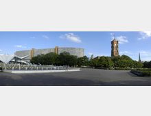 Weitläufiger Platz mit einem Brunnen im Vordergrund, flankiert von Bäumen und Bänken. Im Hintergrund stehen ein modernes Hochhaus, das von dem roten Rathausturm mit Uhr und weiteren Bauwerken überragt wird.