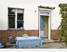 Fassade eines Hauses mit weißem Putz und rotem Klinkersockel, blauer Tür und einem blau gerahmten Fenster. Im Vordergrund steht ein kleiner Gartentisch mit blauer Tischdecke, darauf eine Schale mit Äpfeln, umgeben von passenden blauen Klappstühlen.