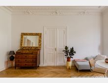 Wohnzimmer mit heller Wandfarbe, verziert mit Stuckarbeiten an der Decke. Im Raum befindet sich eine antike Holzkommode mit vergoldetem Spiegel, eine Pflanze in einem Blumentopf, ein modernes Sofa mit Kissen und eine Sitzgruppe mit einem Tisch und einem geflochtenen Stuhl. Der Boden ist aus hellem Parkett, und eine doppelflügelige weiße Tür prägt die Wand.