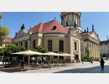 Historisches Gebäude mit rotem Walmdach und Säulenfassade, umgeben von Bäumen; im Vordergrund ein Außenbereich mit Tischen und Stühlen unter großen Sonnenschirmen auf einem gepflasterten Platz.