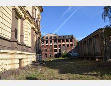 Alt-Text: Eine verlassene Industrieumgebung mit einem historischen, cremefarbenen Gebäude mit verzierten Säulen links im Vordergrund. Im Hintergrund steht ein verfallenes, mehrstöckiges rotes Backsteingebäude mit zerstörten Fenstern. Rechts im Bild befindet sich ein moderneres, schlichtes Betongebäude. Der Boden ist überwuchert von Gras und Wildpflanzen, unter einem blauen Himmel.