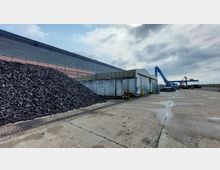 Industrieanlage mit einem großen Haufen von schwarzen Materialien im Vordergrund, einer Wellblechhalle in der Mitte und einem blauen Kran auf einer Betonfläche im Hintergrund. Der Himmel ist bewölkt, und die Umgebung wirkt wie ein Hafengelände oder eine Lagerfläche.