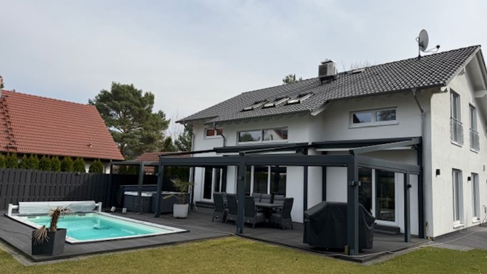 Rear view of a modern two-story white house with a dark tiled roof and a covered patio supported by black posts. A rectangular in-ground swimming pool sits beside a lawn, bordered by decking and a dark privacy fence, with neighboring houses and trees in the background.