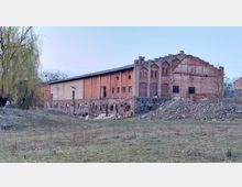 Ein altes, langgezogenes Ziegelgebäude mit historischer, teilweise verfallener Fassade. Der Unterbau besteht aus Natursteinen, das Gebäude steht in einer grasbewachsenen, leicht hügeligen Landschaft mit einem Baum auf der linken Seite.