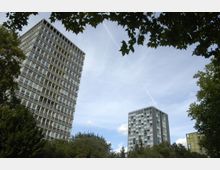 Hochhäuser mit modernen Fassaden, umgeben von Bäumen und blauen Himmel; im Vordergrund dichter Baumbestand, der die Umgebung eines Wohngebiets andeutet.