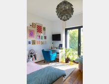 Ein modernes Schlafzimmer mit großen Fenstern, die Blick ins Grüne erlauben. Der Raum enthält ein blaues Samt-Sessel, einen Schreibtisch mit Dekorationen, gerahmte Kunstwerke an der Wand, ein graues Bett sowie eine auffällige Hängelampe aus Metall.