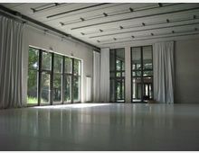 Großer, moderner Raum mit glattem Boden, großen Fensterfronten und weißen Vorhängen, durch die grünes Außenlicht hereinscheint. Die Decke ist mit sichtbaren Metallleisten ausgestattet.