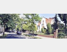 Wohnstraße mit Einfamilienhäusern, gesäumt von Bäumen und geparkten Autos. Im Vordergrund ein weißes Haus mit rotem Ziegeldach, Vorgarten und Zaun.