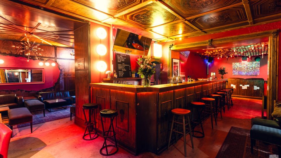 Innenansicht einer Bar mit langer Holztheke und einer Reihe von Barhockern, warm beleuchtet in Rot- und Goldtönen. Auffällig sind die dekorierte Kassettendecke, ein Loungebereich mit Sofas links und ein großer Durchgang zu einem hinteren Raum mit weiterer Theke und Beleuchtung.