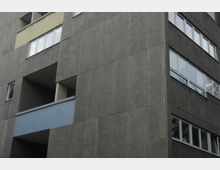 Fassade eines modernen Wohngebäudes mit grauen Betonplatten, großen rechteckigen Fenstern und einem Balkon mit blauer Verkleidung.