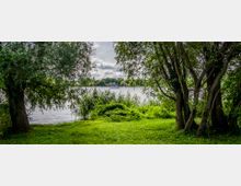 Blick auf einen See durch eine von Bäumen und Sträuchern umrahmte Wiese, mit Gras und dichter Vegetation im Vordergrund und Häusern am gegenüberliegenden Ufer. Im Hintergrund ist ein Boot auf dem Wasser zu sehen, darüber ein bewölkter Himmel.