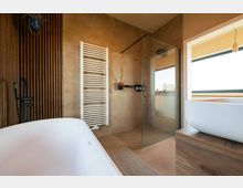 Modernes Badezimmer mit freistehender weißer Badewanne, Glasdusche, Holzpaneelwand, weißem Handtuchheizkörper und einem Waschtisch mit Aufsatzbecken. Ein großes Fenster sorgt für natürliches Licht.