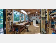 Eine helle Werkstatt mit Holzwänden und zahlreichen Werkzeugen, die an den Wänden und in Regalen organisiert sind. Im Vordergrund steht eine Werkbank mit Hocker, im Hintergrund sind ein Sofa und Lagermöglichkeiten zu sehen; durch die Fenster fällt Licht auf den Raum.