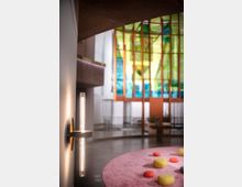 Moderne Innenraumgestaltung mit einer schmalen Wandlampe links im Vordergrund. Im Hintergrund befinden sich farbenfrohe, gläserne Wandpaneele, eine rosa runde Teppichfläche und bunte Sitzkissen, sowie eine helle, offene Raumarchitektur.