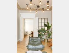 Wohnzimmer mit Holzfußboden, Sessel aus grünem Leder, großer Zimmerpflanze und modernem Kronleuchter an der Decke. Im Hintergrund offene weiße Türen, die in einen weiteren Raum mit heller Küche und Schreibtisch führen.