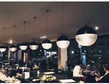 Ein modernes Restaurant mit einer langen Theke im Vordergrund, bestückt mit Getränkezubehör und Obstschalen. Mehrere runde Pendelleuchten hängen von der Decke, während Gäste an Tischen vor großen Fenstern sitzen, durch die eine nächtliche Straßenszene sichtbar ist.