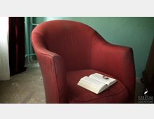 Roter gepolsterter Sessel mit einem offenen Buch und einer Uhr darauf, vor einer mintgrünen Wand; im Hintergrund ein weißer Vorhang und ein rotbrauner Vorhang.