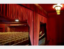 Theaterraum mit roten Vorhängen und warm beleuchteten Wänden, gepolsterte beige Sitzreihen in einem Zuschauerraum und eine kleine Loge im Vordergrund, beleuchtet von einer Deckenleuchte.