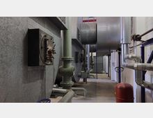 Industrieanlage mit großen metallischen Lüftungskanälen, Rohren, Ventilen und technischen Komponenten in einem hell beleuchteten Innenraum. Der Boden ist aus glattem Beton, und die Wände bestehen aus weißen Ziegeln und Metallverkleidungen.