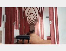 Nördliches Seitenschiff einer gotischen Kirche mit roten Spitzbögen und weißen Wänden. Im Vordergrund steht ein schwarzer Flügel auf einem Boden aus roten Fliesen.