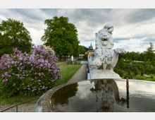 Steinlöwenskulptur auf einem Podest in einem Park, umgeben von blühenden lila Fliederbüschen und grünen Rasenflächen; im Hintergrund befinden sich Bäume, eine historische Gebäudeuhr und eine Statue. Im Vordergrund spiegelt sich der Himmel in einem flachen Brunnen.