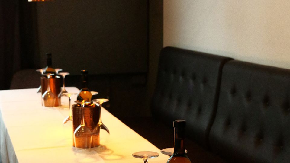 Langer, schmaler Restauranttisch mit weißer Tischdecke, gedeckt mit umgedrehten Weingläsern und Flaschen in Metallkühlern. Darüber hängen mehrere kupferfarbene Pendelleuchten, daneben verläuft eine dunkel gepolsterte Sitzbank entlang der Wand mit einem Wandbild.