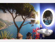 Wand eines Zimmers mit einer großen Fototapete, die eine mediterrane Küstenlandschaft mit Bäumen, Meer und Architektur zeigt. Rechts befindet sich ein beleuchteter ovaler Spiegel über einem weißen Waschbecken, mit einem Seifenspender auf einem kleinen Regal.