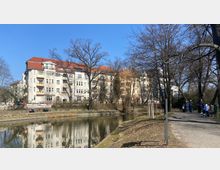 Wohngebäude im Stil des frühen 20. Jahrhunderts mit roten Ziegeldächern und hellen Fassaden, am Ufer eines ruhigen Kanals. Im Vordergrund ein Gehweg mit Spaziergängern, Bänken und Bäumen ohne Laub.