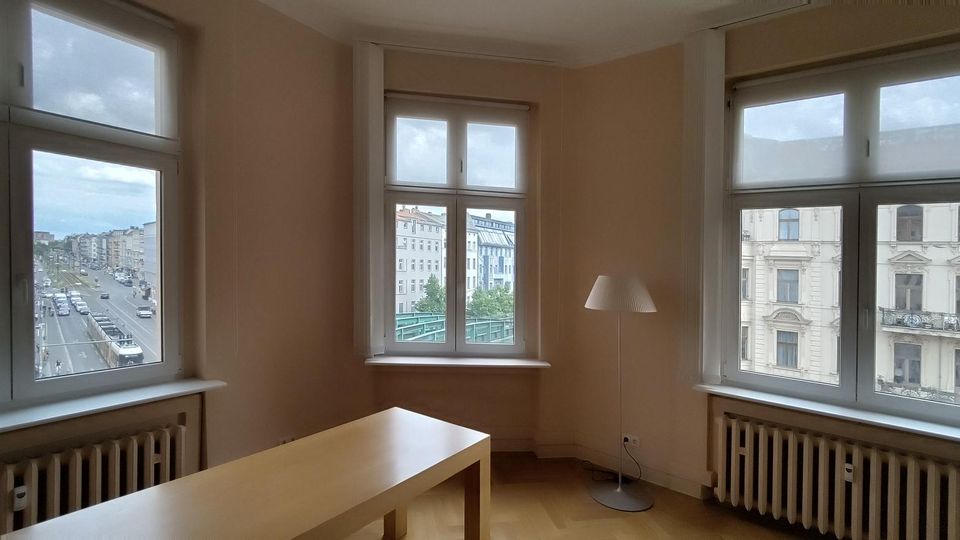Heller, leerer Wohnraum mit hellen Wänden, Holzfußboden und großformatigen Eckfenstern auf zwei Seiten sowie einem weiteren Fenster an der Stirnseite. Vor den Fenstern stehen Heizkörper; im Raum befinden sich ein schlichter Holztisch und eine Stehlampe, mit Blick auf eine städtische Straße und gegenüberliegende Altbaufassaden.