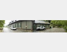 Panoramaaufnahme eines historischen Gebäudes aus Holz mit grün gestrichenen Fensterläden und einem Schieferdach. Im Vordergrund sind gepflasterte Wege zu sehen, links ein weißes Auto, rechts ein silbernes SUV; umgeben von Bäumen und Grünflächen.