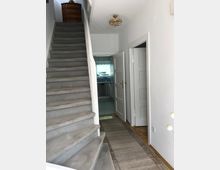 Flur in einem Wohnhaus mit einer Teppichtreppe, die nach oben führt, und einer Kommode auf dem Treppenabsatz. Eine offene Tür führt in eine helle Küche, und eine Hängelampe mit Kristallelementen ist an der Decke angebracht.