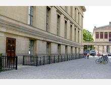 Straßenszene mit einem historischen Gebäude aus beigefarbenem Stein rechts im Bild, das durch große Fenster und dezente Ornamentik gekennzeichnet ist. Im Hintergrund sind weitere klassische Gebäude, Menschen sowie geparkte Fahrräder und Fahrzeuge zu sehen.