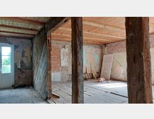 Ein unfertiger Innenraum mit freiliegenden Holzbalken und unverputzten Backstein- und Putzwänden. Der Boden besteht aus alten Holzdielen, und an einer Wand lehnt eine weiße Tür; im Hintergrund ist eine Glastür, die nach außen führt.