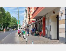 Straßenszene in einer Stadt mit Wohngebäuden und Geschäften, teils mit rot-beigen Fassaden. Am Bürgersteig stehen Tische und Stühle, daneben sind Fahrräder, E-Scooter und geparkte Autos; eine Straßenbahn fährt im Hintergrund auf einer von Bäumen gesäumten Straße.