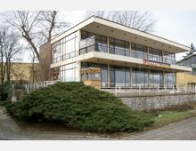 Modernes zweistöckiges Gebäude mit großen Glasfassaden und Flachdach, beschriftet mit "Champions Teehaus". Im Vordergrund befinden sich eine Treppe, ein gepflasterter Gehweg und niedrige Büsche.