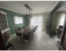 Esszimmer mit einem langen Holztisch und mehreren gepolsterten Stühlen, grüne Wände, ein großer Wandspiegel, ein hängender Kronleuchter sowie eine weiße Anrichte. Im Hintergrund führen bodentiefe Fenster mit transparenten Vorhängen zu einer Terrasse.