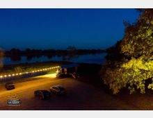 Nachtaufnahme eines ländlichen Geländes mit einem beleuchteten Anhänger, einer Lichterkette und parkenden Autos auf einem gepflasterten Platz. Im Hintergrund ist ein Fluss mit dunklen Uferbäumen sichtbar, rechts im Bild ein beleuchteter Baum.