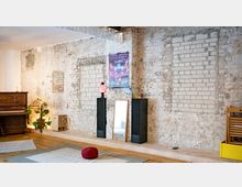 Innenraum mit rohen Backstein- und Putzwänden, in denen mehrere zugemauerte Fensteröffnungen sichtbar sind, sowie Holzboden und Deckenbeleuchtung. An der Wand stehen zwei hohe schwarze Lautsprecher und ein Spiegel; links sind ein Klavier und Pflanzen zu sehen.