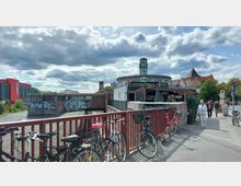 Straßenszene in einer städtischen Umgebung mit einer roten Fußgängerbrücke im Vordergrund, an der mehrere Fahrräder angelehnt sind. Im Hintergrund ein S-Bahn-Schild auf einem runden Gebäude mit der Aufschrift „Döner Express“ und eine Ziegelmauer mit Graffiti; daneben Wohn- und Gewerbegebäude sowie Gehwege mit Fußgängern.