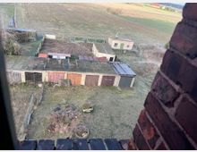 Blick aus einem Gebäude auf eine Reihe alter Garagen mit teils verwitterten Türen sowie ein angrenzendes umzäuntes Gelände mit Schuppen und einem kleinen Gebäude. Im Hintergrund erstrecken sich Felder und Wiesen.