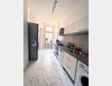 Schmale, helle Küche mit weißer Einrichtung, Marmorfliesenboden und großen Fenstern im Hintergrund. Auf der rechten Seite befinden sich Arbeitsflächen, ein Spülbecken und ein Einbauherd, links ein großer silberner Kühlschrank.