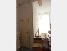 Ein kleines Badezimmer mit einer weißen Badewanne unter einem großen Fenster, das durch einen halbtransparenten Vorhang bedeckt ist. Links befindet sich eine schlichte Holztür, und rechts stehen ein Regal und ein kleiner Tisch mit Handtüchern und Pflegeartikeln.