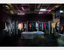 Ein moderner Bekleidungsladen mit dunklen Wänden, einer offenen Decke mit sichtbaren Lüftungselementen sowie fluoreszierender Beleuchtung. An Gitterständern hängen farbenfrohe Kleidungsstücke, und in der Mitte befinden sich niedrige Tische aus Metallgitter.